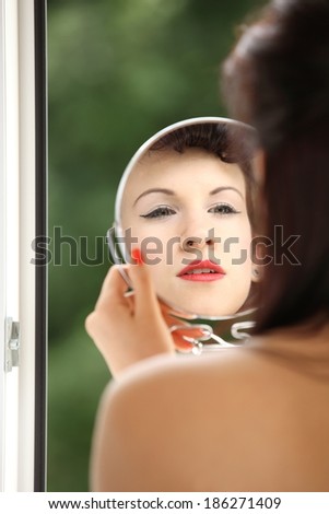 Young woman preparing to party indoor, girl retro style applying make up red lipstick looking at mirror green blurred background