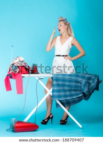 Full length sexy girl retro style ironing male shirt, tired woman housewife in domestic role. Traditional sharing household chores.  Pin up housework.  Vivid blue background