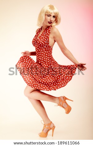 Vintage pinup style. Full length of stylish woman dancing. Girl in blond wig and retro spotted red dress on pink. Disguise. Party.