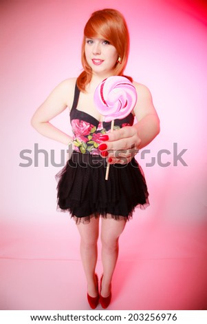 Full length of sexy young woman showing giving candy. Redhair girl holding sweet lollipop on pink. Studio shot. Sweets.