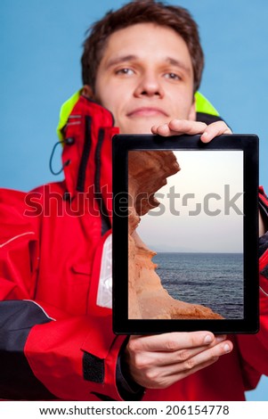 Man in red wind jacket holding ipad with photo of sea seascape. Traveler showing screen tablet touchpad dreaming about summer holiday. Technology and travel.