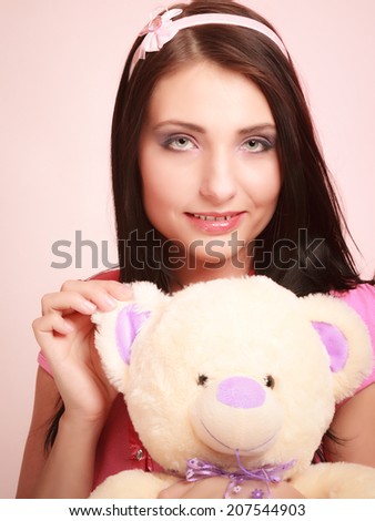 Portrait of childish young woman with headband holding toy. Infantile girl hugging teddy bear on pink. Longing for childhood. Studio shot.