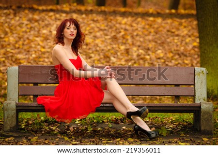 Beautiful young woman in elegant red dress sitting on bench. Girl relaxing on autumnal park. Fall leisure. Outside.