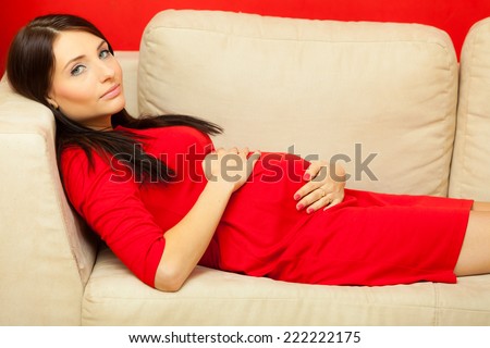 Pregnancy, motherhood and happiness concept. Beautiful sexy stylish elegant pregnant woman in red dress relaxing on sofa and touching her belly