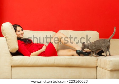 Pregnancy, motherhood and happiness concept. Beautiful elegant pregnant woman in red dress relaxing on sofa playing with cat pet.
