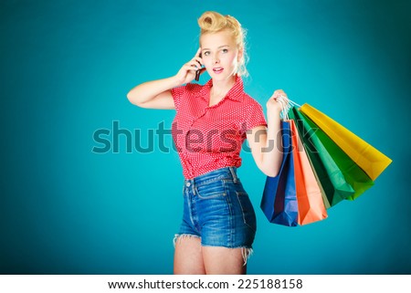Pinup blond girl young woman in retro style calling on the mobile phone. Client customer holding colorful paper shopping bags on vibrant blue. Retail and sale. Studio shot.