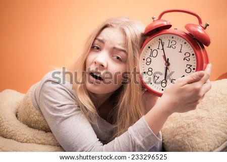 Not want to wake up concept. Funny young woman in bed waking up late. Unhappy sleepy girl holds red alarm clock.