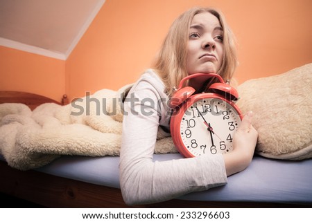 Not want to wake up concept. Funny young woman in bed waking up late. Unhappy sleepy girl holds red alarm clock.