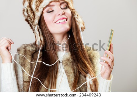 young beautiful woman with smart phone listening music in earphonees. Happy girl in winter clothes fur cap dancing on gray