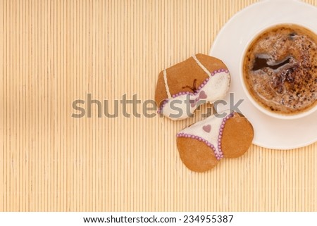 White cup of coffe with funny bikini underwear shape gingerbread cake cookie sweet dessert with white icing and violet decoration border or frame on beige bamboo mat background