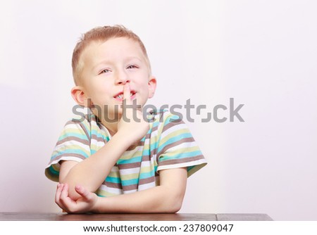 Portrait of blond pensive thoughtful boy or child kid asking for quiet with finger on lips at the table interior. Emotions. Copyspace.
