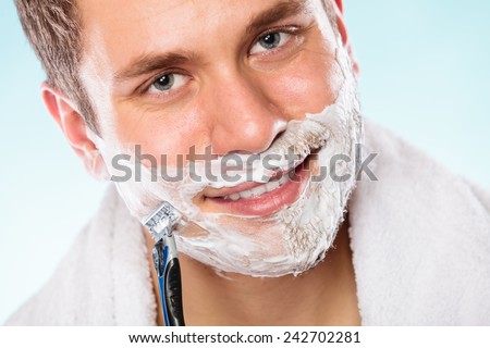 Health beauty and skin care concept. Handsome young bearded man with foam on face shaving with razor on blue background.