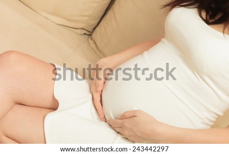 Pregnancy, motherhood and happiness concept. Beautiful stylish elegant pregnant woman in white short dress relaxing on sofa touching her belly, at home