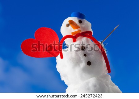 Little happy christmas snowman in blue screw top as hat red scarf and with clip red heart love symbol toothpick outdoor. Winter season seasonal specific valentine\'s day. Blue sky background.
