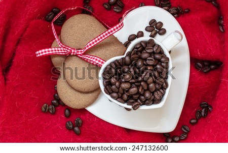 Coffee love concept. Coffee beans in heart shaped white cup and sweet cookie gingerbread with ribbon on red cloth background. Top view