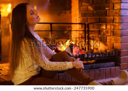 Winter at home, heating. Attractive woman long hair girl closed eyes relaxing warming up at fire fireplace. Indoor.