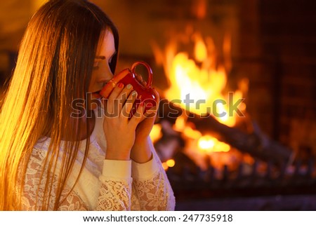 Winter at home, heating. Closeup woman face, smiling long hair girl relaxing warming up at fire fireplace, holding mug with hot drink. Indoor.