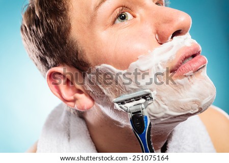 Health beauty and skin care concept. Handsome young bearded man with foam on face shaving with razor on blue background. Unusual wide angle view