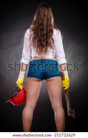Woman in male specialities. Safe by work. Feminist sexy woman with tools hatchet and red helmet Back view on black grey background.