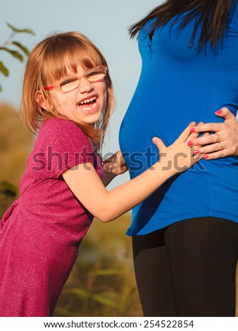 Pregnancy, motherhood and happiness concept. Pregnant woman walking outdoor in park, her daughter happy little girl holding belly of mom
