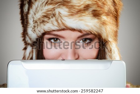 Technology internet concept. Closeup fashion woman in winter clothes fur cap covering her face with tablet computer studio shot on gray