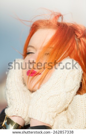 Winter fashion. Beauty redhaired head with hair blowing on wind, woman in warm clothing outdoor enjoying sunlight.