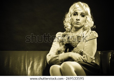 Mental disorder concept. Young childlike woman wearing like puppet doll sitting with teddy bear toy on red couch dark black background
