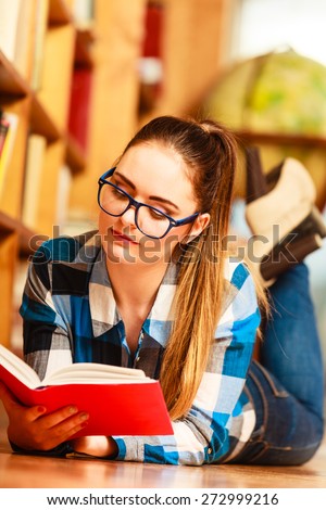 Education school concept. Casual female student fashionable girl in blue glasses lying on floor in college library with book reading. Indoor