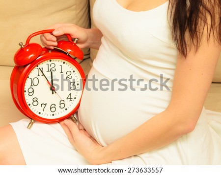 Pregnant woman holding red alarm clock waiting, sitting on sofa at home