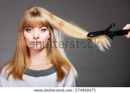 Hairstyling. Attractive blonde woman long haired making hairstyle hairdo with electric hair iron straightener gray background