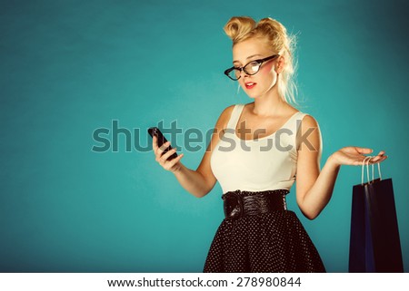 Retro style. Young woman pinup girl in glasses with shopping bag using cell phone. Blue background.