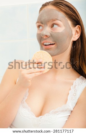 Beauty procedures spa and skin care concept. Young woman with facial clay mask in bathroom holds sponge to remove mud