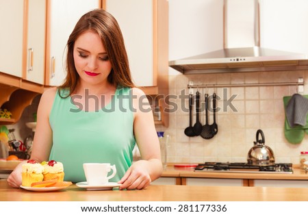 Woman with cup of coffee and delicious gourmet sweet cream cake cupcake and orange. Glutton girl sitting in kitchen with hot beverage having breakfast. Appetite and gluttony concept.