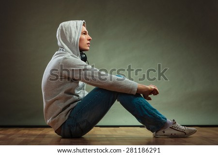 Music passion youth concept. Serious hooded man teen boy with headphones sitting daydreaming on floor grunge background