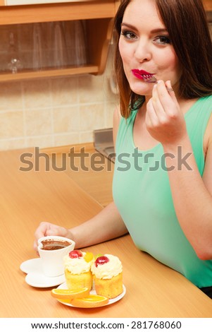 Woman with cup of coffee eating delicious gourmet sweet cream cake cupcake and orange. Glutton girl sitting in kitchen with hot beverage having breakfast. Appetite and gluttony concept.