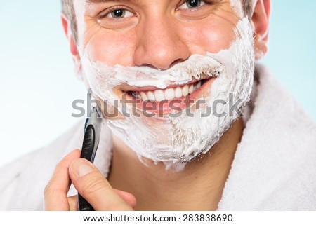 Health beauty and skin care concept. Handsome young bearded man with foam on face shaving with razor on blue background.