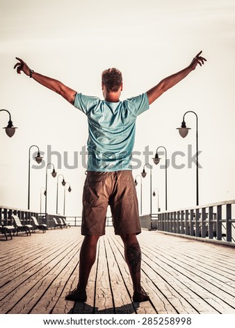 Man tourist on pier with raised hands arms. Guy enjoying summer travel vacation by sea. Fashion. Happiness and freedom concept.