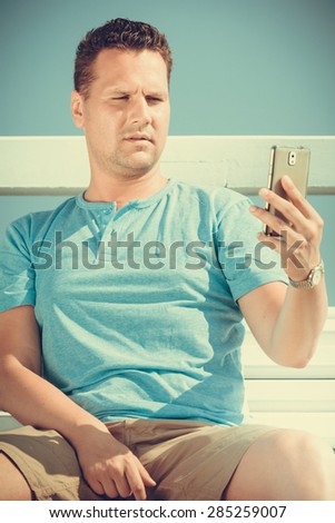 Handsome man tourist sitting on bench on pier using smartphone. Fashion guy enjoying summer travel vacation by sea. Relax and technology concept.
