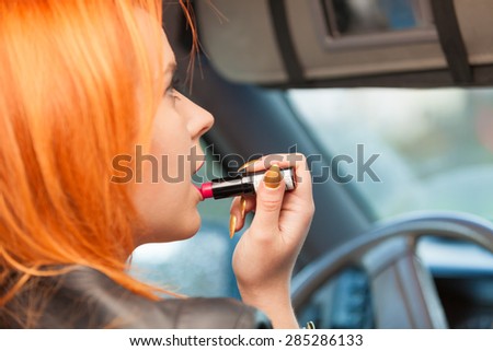 Concept of danger driving. Young woman driver red haired teenage girl painting her lips doing applying make up while driving the car.