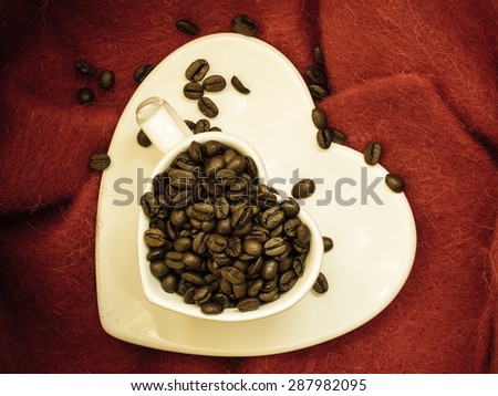 Coffee klatsch java concept. Heart shaped white cup filled with roasted coffee beans on red cloth background