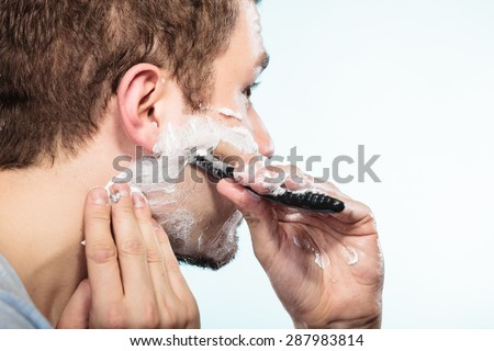Health beauty and skin care concept. Closeup young bearded man with foam on face shaving with razor on blue background.