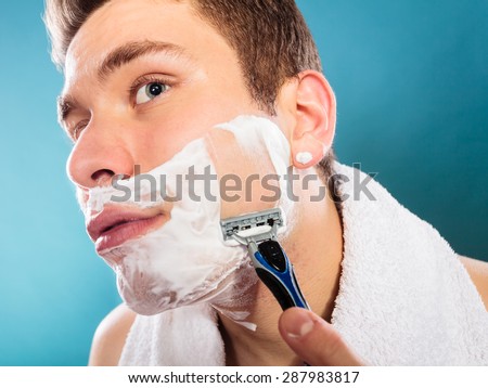 Health beauty and skin care concept. Handsome young bearded man with foam on face shaving with razor on blue background. Unusual wide angle view