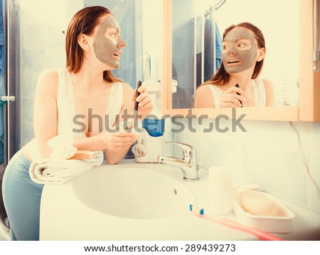 Beauty skin care cosmetics and health concept. Young woman holding brush and bowl with facial clay mask applying mud to her face in bathroom