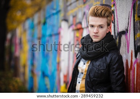 Portrait of handsome trendy man outdoor in city setting, male model wearing winter clothes black jacket and scarf against colorful graffiti wall