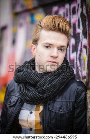 handsome trendy man outdoor in city setting, male model wearing black jacket scarf and checked shirt against colorful graffiti wall