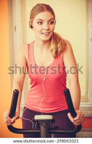 Active young woman working out on exercise bike stationary bicycle. Sporty girl training at home listening music. Fitness and weight loss concept. Instagram filtered.