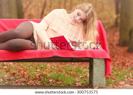 Happy smiling woman relaxing in fall park reading book. Young blonde girl resting laying on bench. Autumn lifestyle fun.