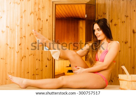 Young woman in wood finnish spa sauna massaging skin with exfoliating glove. Girl in bikini relaxing. Skincare concept.