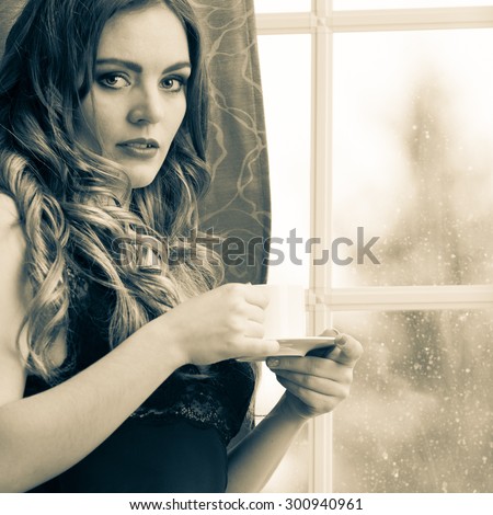 Sensual seductive woman in lingerie drinking cup of coffee by curtain and french door window at home. Young girl with hot energizing beverage stay awake. Caffeine energy. Black and white.