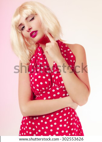 Vintage pinup style. Portrait of beautiful stylized thoughtful young woman winking. Attractive pensive girl in blond wig and retro spotted red dress on pink. Disguise. Studio shot.